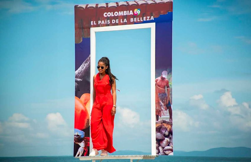 Mujer morena, con enterizo rojo y gafas de sol, posando para una foto en medio de la playa y de fondo el mar.